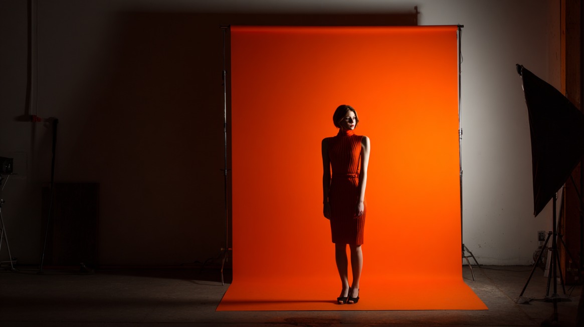 Woman in a red dress standing in front of a vibrant orange backdrop with studio lights set up around her